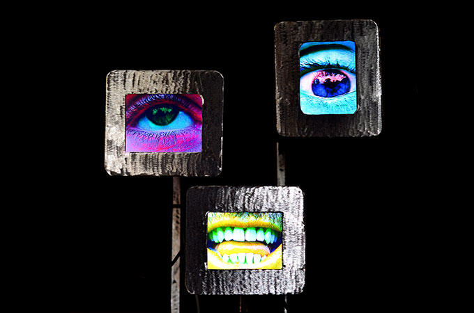 Abstract artwork of a robot with three screens – one with picture of a pink eye, one with picture of a blue eye and one with picture of a yellow mouth on a black background