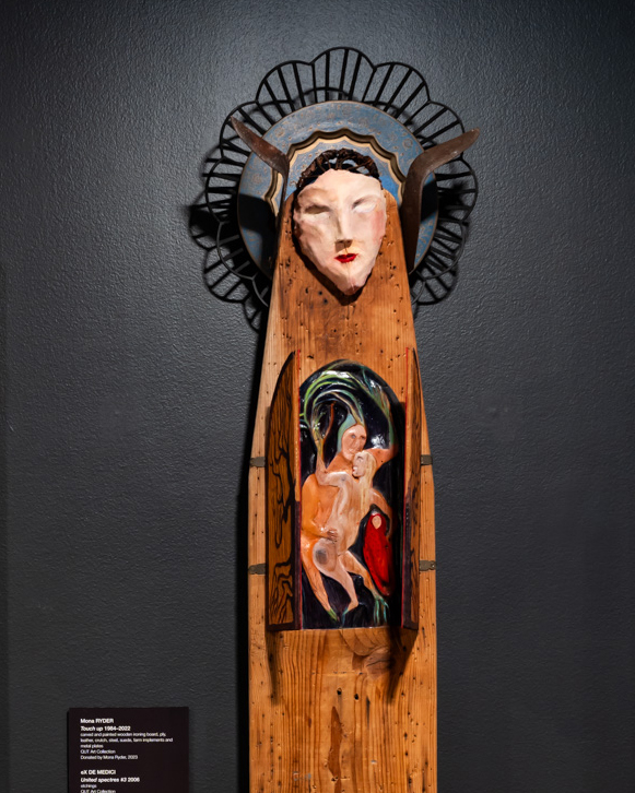 A carved and painted wooden ironing board mounted vertically on the wall, with a mask of a woman's face at the top